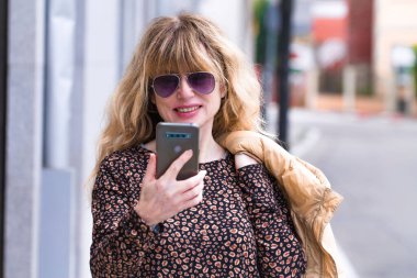 Güneş gözlüklü ve ceketli gülümseyen kadın sokakta telefon kullanıyor.