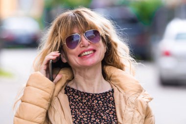 Güneş gözlüklü ve ceketli gülümseyen kadın sokakta telefon kullanıyor.