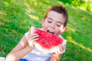 Genç bir çocuk sahilde karpuz yiyor.