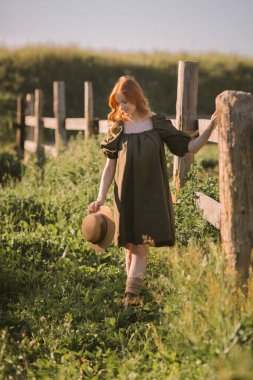 Kızıl saçlı bir kız tahtadan yapılmış büyük bir çitin yanında yürüyor..