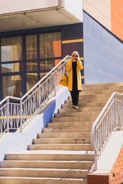 Sarı saçlı, parlak sarı ceketli ve güneş gözlüklü zarif modern kadın şehir caddesinde yürüyor. Şehir tarzı modern kadın alışverişi.
