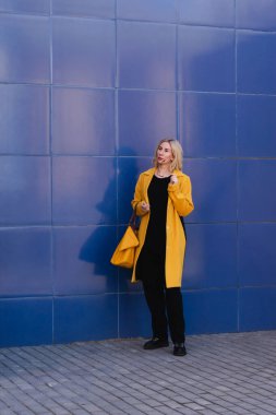 Sarı saçlı, parlak sarı ceketli, mavi arka planda, şehir caddesinde yaşayan genç bir kadın. Şehir tarzı modern kadın.