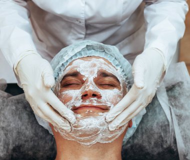 Beautician applying scrub mask on face to young man, spa beauty salon. The concept of body and face care for men