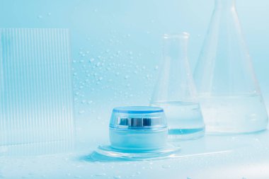 Kavanozda kozmetik kremi, mavi arka planda cam mataralarla su sıçratır ve damlatır.