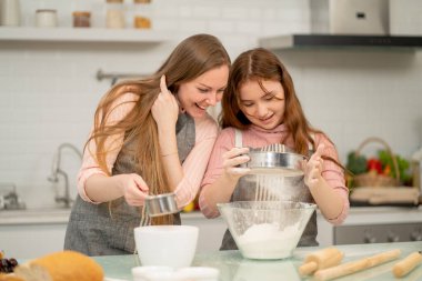 Önlüklü beyaz bir anne kızını un ayıklayarak kek pişirme sürecine teşvik ediyor ve birlikte mutfaktan mutluluk ve neşeyle keyif alıyor..