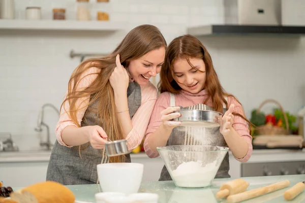 Önlüklü beyaz bir anne kızını un ayıklayarak kek pişirme sürecine teşvik ediyor ve birlikte mutfaktan mutluluk ve neşeyle keyif alıyor..