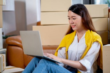 Genç Asyalı kadın, internet üzerinden müşteriye ürün dağıtımı için dizüstü bilgisayar kullanıyor ve küçük işletme ve yeni modern satış ve alışveriş tarzı konseptiyle işyerinde kalıyor.