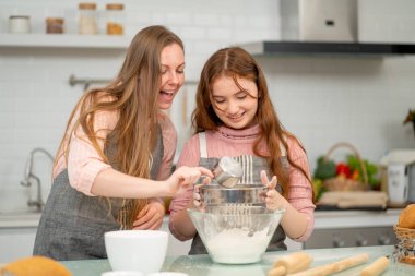 Önlüklü beyaz bir anne kızını un ayıklayarak kek pişirme sürecine teşvik ediyor ve birlikte mutfaktan mutluluk ve neşeyle keyif alıyor..