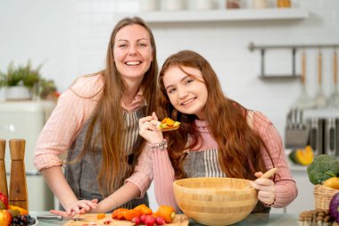 Genç kız karışık sebzelerle kaşık tutuyor ve kameraya gülümsüyor ve annesiyle mutfakta kalıyor. Mutluluk ve aile aktivitelerinin keyfini çıkarıyor gibi görünüyor..