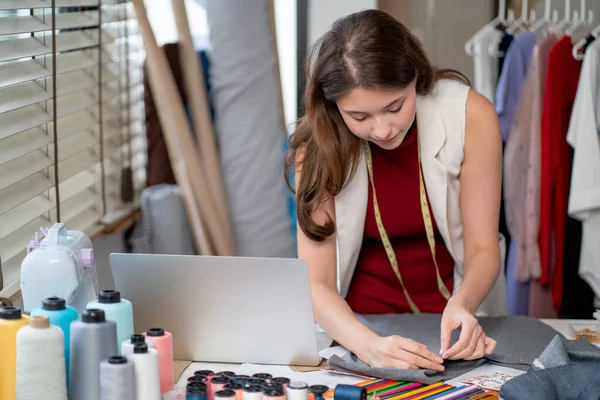 Genç tasarımcı veya güzel terzi kadın dizüstü bilgisayarın önünde çalışır ve yeni modern stile sahip işyeri alanındaki müşteriler için kumaş tasarımı için araçlar veya aksesuarlar kullanır..