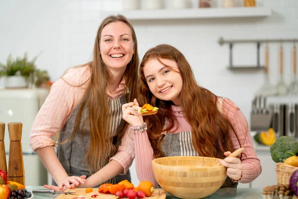 Genç kız karışık sebzelerle kaşık tutuyor ve kameraya gülümsüyor ve annesiyle mutfakta kalıyor. Mutluluk ve aile aktivitelerinin keyfini çıkarıyor gibi görünüyor..