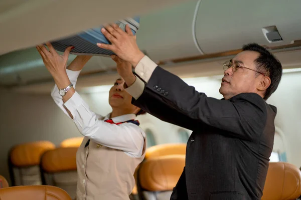 Business man bring his luggage to overhead shelf in airplane and air hostess help and support him during go to work in other city.