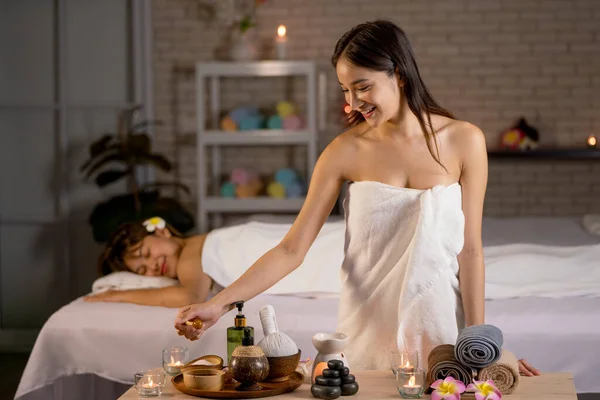 Asian beautiful woman wearing a towel stand in front of other woman lie on bed and she hold stick with honey during process of massage and spa.
