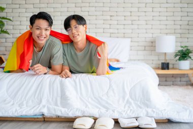 Gay ya da Igbt 'li bir çift beyaz yatağın üzerinde gökkuşağı bayrağı kullanırlar. Yatak odasında gülümseyen kameraya da bakarlar ve mutlu görünürler..