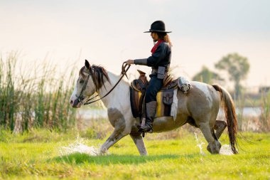 Şapka kontrol atı olan bir kovboy nehir kenarındaki çim tarlasında yürüyor ve yürürken biraz su sıçratıyor..
