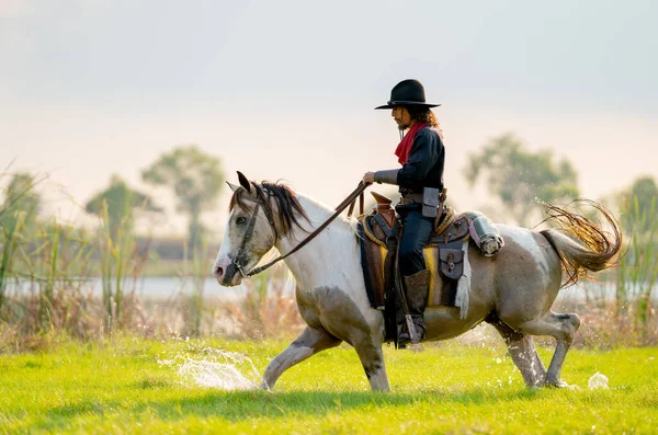 One cowboy with hat control horse to walk through grass field cover by water near river and show some splash during walking.