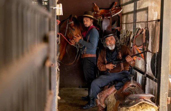 Two man with cowboy costume stay in horse stable that senior check long gun and other one take care horse in the back and they look at camera.