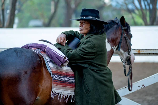 Asian old man with cowboy costume stand and lean beside of dark brown horse and look to the left side and stay near outdoor stable wait to train the horse.