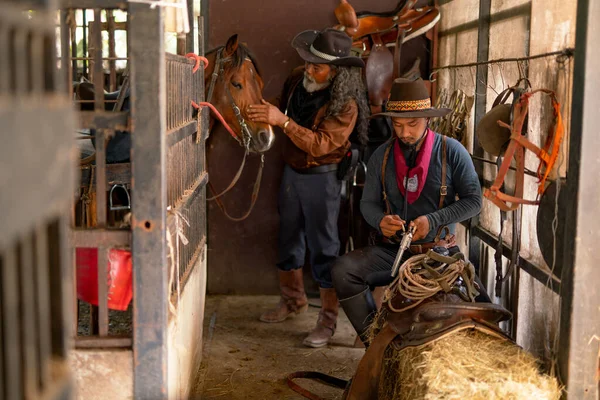 Two man with cowboy costume stay in horse stable that one check the bullet of short gun and other one take care horse in the back.