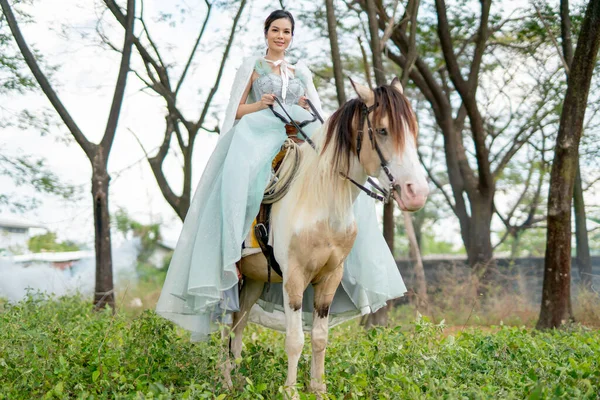 Beautiful woman with blue dress stay on white horse and stand in field near village during early morning also look at camera.