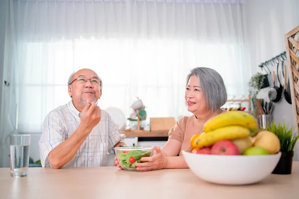 Asian senior or elderly man and woman enjoy with vegetable salad together during stay in kitchen in their house with various types of fruits on the table.