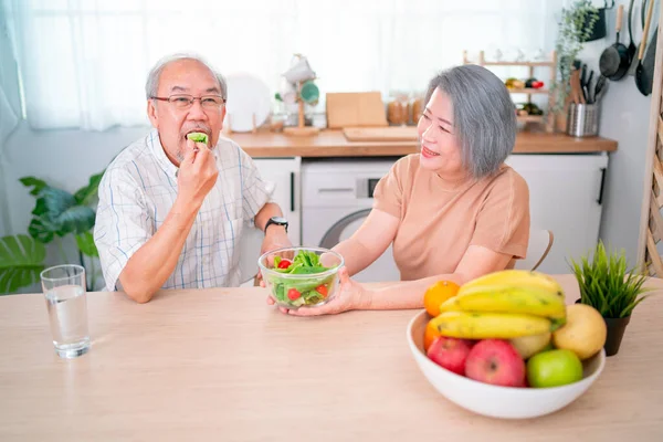 Asian senior or elderly man and woman enjoy with vegetable salad together during stay in kitchen in their house with various types of fruits on the table.