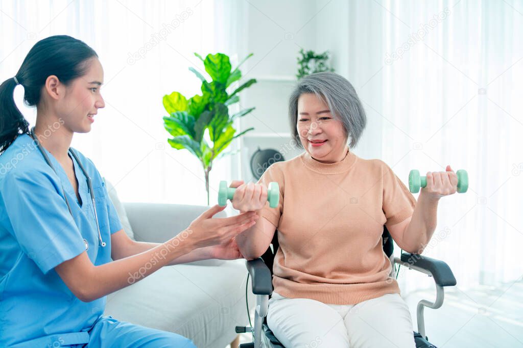 Asian nurse or doctor who work as homecare support staff help senior woman exercise with using dumbbell and stay in living room.