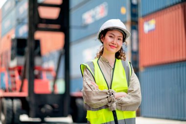 Asyalı kargo konteynırı işçisi veya teknisyeninin portresi kolları çaprazlanmış duruyor ve iş yerindeki kameraya gülümsüyor..