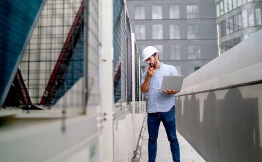 Profesyonel mühendis ya da teknisyen elinde dizüstü bilgisayarla ayakta duruyor ve inşaat alanının terasındaki makinenin yanındaki yolda telsiz bağlantısı kuruyor ve ekipmanların işlevlerini kontrol ediyor..