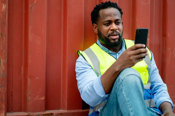 African American cargo container worker sit or lean to container tank also use mobile phone to contact coworker with happiness.