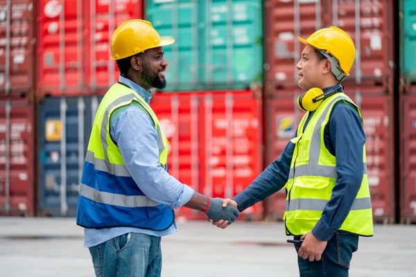 African American worker or technician shake hands with Asian staff in cargo container workplace area with stack of container tank as background.