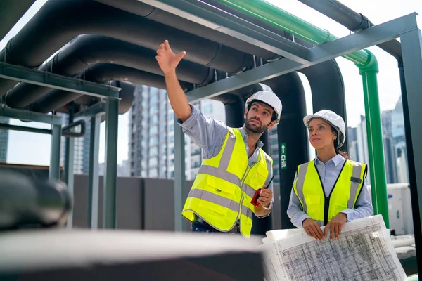 Two professional engineer discuss with blue print of construction site and stay under the floating pipe or pipe hanging on deck of building.