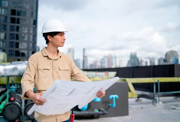 Asian engineer with white safety helmet hold and look on the right and hold blue print paper of construction site also stay on terrace or deck of the building.