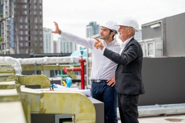 Young engineer point his hand to the front and explain to senior coworker and they also stay near line of air vents at construction site.