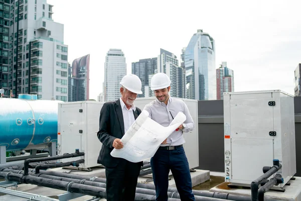 Wide shot of two engineers or technician man discuss together using building plan or drawing and they also stay on rooftop area of construction site.