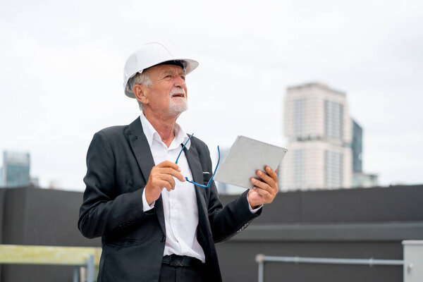 Senior engineer man using tablet to check and maintenance in area of construction site with other building as background.