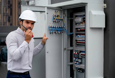 Professional engineer man open lid of electrical control panel and use walkie talkie to contact and ask for support from his coworker at workplace of construction site.