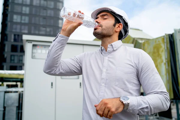 Engineer man or technician worker drink water from bottle and stand in front of electrical control panel of construction site.