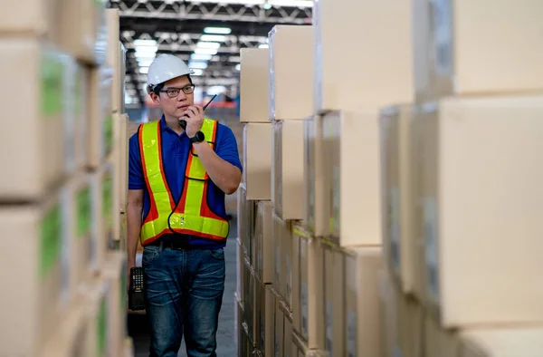 Asian warehouse worker or manager use walkie talkie to contact his team and stand between stack of product carton.
