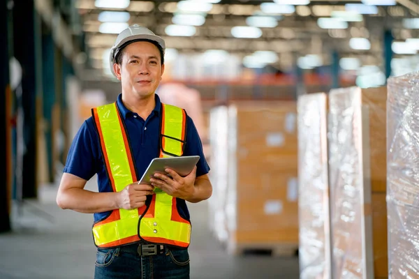 Portrait of Asian warehouse worker or manager hold tablet and look at camer also stand in front of stack of product carton. Concept of professional factory worker support for delivery  transport.