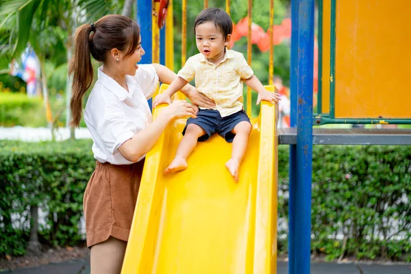 Asian mother stand and take care little boy play slide in fun park of garden and they look happy to have enjoy time together.