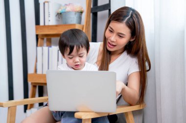 Asian mother enjoy to have fun with using laptop with her son and sit in front of white curtain and day light and they look happy together.