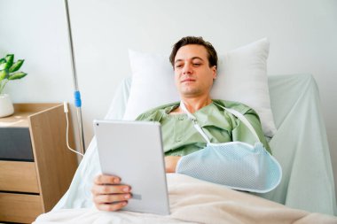Kafkasyalı hasta yatağa yatar ve kol tedavisi sonrası rahatlamak için tablet kullanır ve hasta odasında kalır..