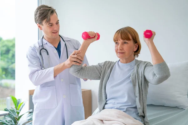 Caucasian doctor support senior patient woman who sit on bed and lift up dumbbell to exercise after treatment in hospital.