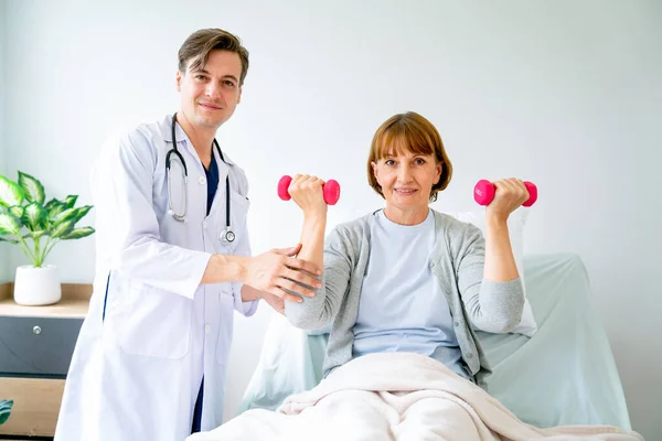 Caucasian doctor support senior patient woman who sit on bed and lift up dumbbell to exercise after treatment in hospital. They also look at camera with concept of elderly healthcare for healthy life.