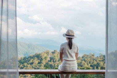 Happy tourist woman relaxing and looking mountain view at countryside home or homestay in the morning. Vacation, blogger, SoloTravel, journey, trip and relaxing concept