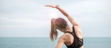 Spor kıyafetli genç bir kadın, vücudu okyanus manzarasına karşı esnetiyor, sabahları sağlıklı kadın egzersizi yapıyor. Spor, sağlık ve iş hayatı dengesi kavramları