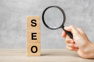 SEO (Arama Motoru İyimserleştirme) metin tahta küp blokları ve masanın üzerinde el ele tutuşan büyüteç. Fikir, Strateji, Reklam, Pazarlama, Anahtar Kelime ve İçerik kavramı