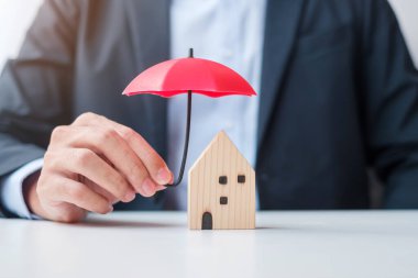 İşadamı elinde kırmızı Şemsiye ile tahta ev modelini tutuyor. Gayrimenkul, sigorta ve mülkiyet kavramları
