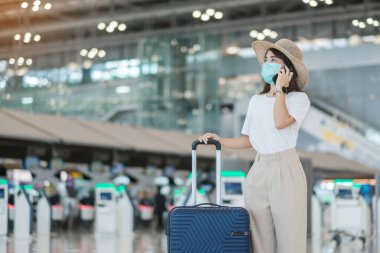 Yüz maskesi takan ve havaalanında cep telefonu kullanan Asyalı kadın turist Coronavirus hastalığı enfeksiyonunu koruyor. Aşı artırıcı doz konseptinden sonra seyahat zamanı